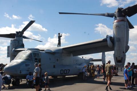 A U.S. Air Force member stands atop a grounded Osprey aircraft as people watch another Osprey in action during the air show.