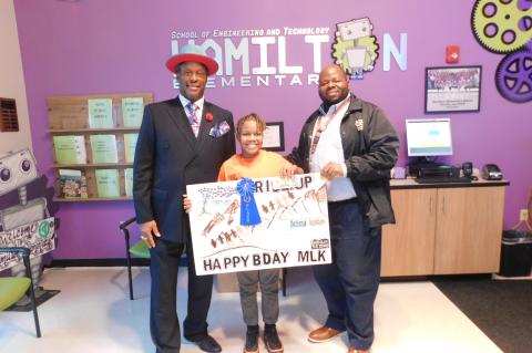 The first place winner of the MLK Jr. Poster Competition is Adrian Fuller III a student at Hamilton Elementary. Adrian received a monetary prize. L-R: Co-Chairman of the MLK Jr. Steering Committee Kenneth Bentley, Adrian Fuller III, Principal of Hamilton Elementary and Adrian’s father, Mr. Adrian Fuller Jr.