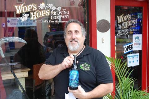 Wop’s Hops Owner Greg Piecora in front of the brewery holding his anniversary brew, VI Fathoms Down.  